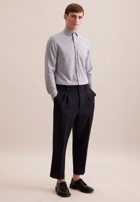 Helles blaues kariertes Hemd mit Kragen, knöpfbarer Front; kombiniert mit dunkelblauen plissierten Hosen und schwarzen Schnürschuhen.