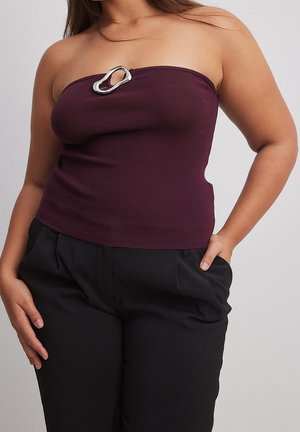 Kobieta ubrana w fioletowy, bezramkowy prążkowany top z srebrną ozdobną sprzączką, do tego czarne spodnie z wysokim stanem, ręka w kieszeni.