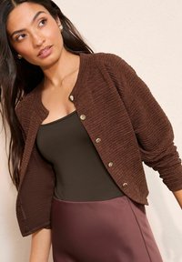 Brązowy sweter kardigan o fakturowanej strukturze, z rękawami do połowy przedramienia, złotymi guzikami oraz okrągłym dekoltem, w połączeniu z ciemnobrązową bluzką i bordową spódnicą.