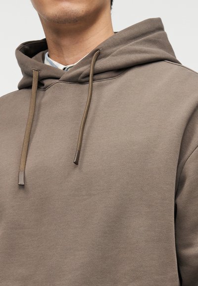 Άνδρας που φοράει ανοιχτό καφέ φούτερ με κουκούλα και κορδόνια πάνω από ανοιχτόχρωμο πουκάμισο με γιακά, κομμένο από το σαγόνι μέχρι το στήθος.