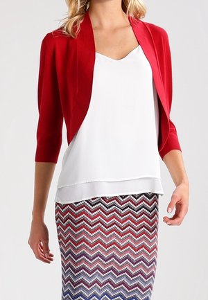 Frau trägt eine rote, bauchfreie Strickjacke, eine weiße ärmellose Bluse und einen hoch taillierten Rock mit Zickzackmuster in Rot, Lila und Weiß.