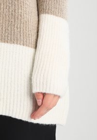 Sweter dzianinowy o prążkowanej teksturze, z beżowymi i białymi poziomymi paskami. Szerokie rękawy mają lekko podwinięty brzeg.