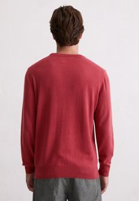 Czerwony sweter z okrągłym dekoltem i długimi rękawami, wykonany z teksturowanej tkaniny, z prążkowaniem na mankietach i u dołu, noszony na szarych spodniach, widziany od tyłu.