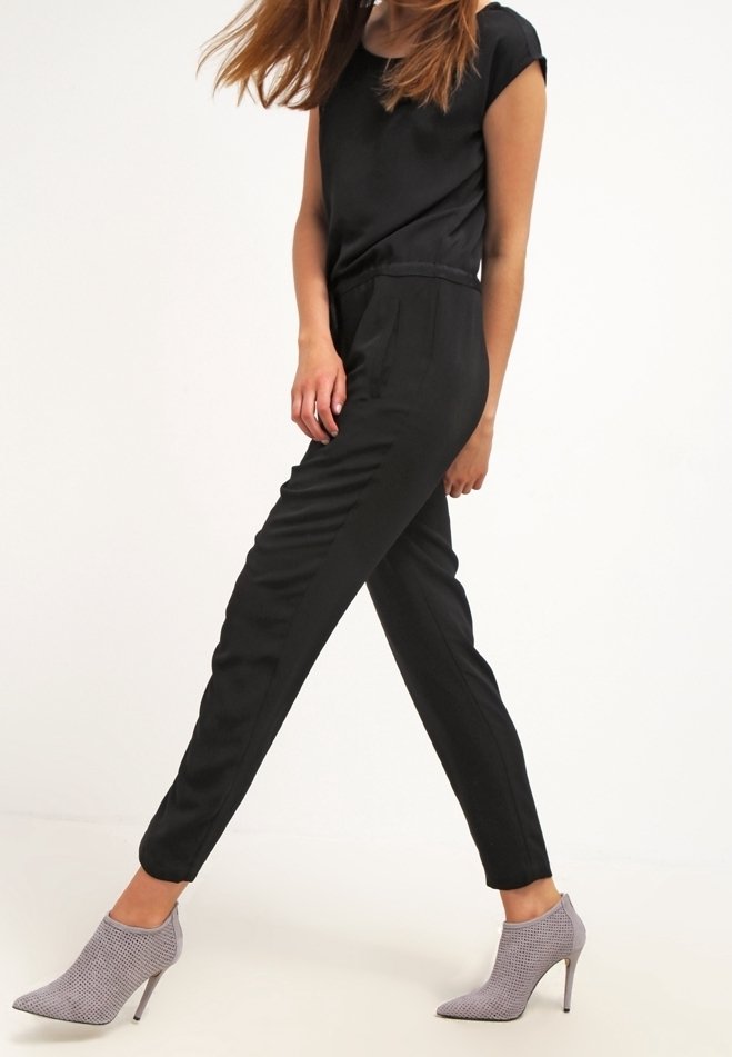 Svart, åtsittande jumpsuit med korta ärmar och avsmalnande ben; har en slät yta. Matchad med ljusgrå, mönstrade högklackade skor.