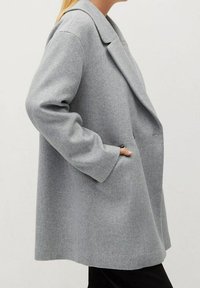 Kobieta w luźnym, jasnoszarym wełnianym płaszczu z szerokimi klapami, z ręką w bocznej kieszeni, widziana z profilu.