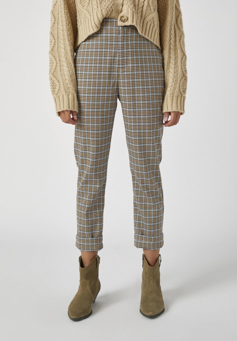 Personne portant un pull beige en maille torsadée, un pantalon à carreaux taille haute retroussé aux chevilles, et des bottines en daim marron clair.