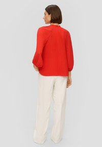 Rode geplooide blouse met lange mouwen en een hoge halslijn, gecombineerd met witte wijde broek. Zachte textuur en levendige kleur.