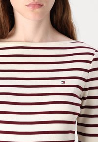 Dryžuotas kreminės ir burgundinės spalvos topas, turintis laivagalio iškirptę ir mažą logotipo ženklelį. Minkštas, prigludęs dizainas su horizontaliais dryžiais.