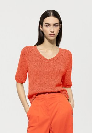 Gestrickter, orangefarbener Pullover mit V-Ausschnitt und kurzen, leicht gepufften Ärmeln, kombiniert mit passenden orangen Hosen. Die Textur wirkt weich und leicht.