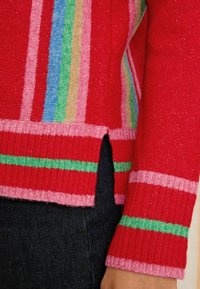 Piros pulóver színes csíkokkal, rózsaszín bordázott mandzsettákkal és hem, valamint oldalsó hasítékokkal. Puha textúrájú, enyhén fényes felülettel.