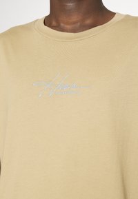 Beige bavlněné tričko s kulatým výstřihem, na levé části hrudi s vyšitým světle modrým textem "Hollister CALIFORNIA".