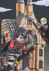 LEGO-tema T-skjorte som viser to figurer som flyr på sopelimer foran et detaljert slott med grå, rød og beige elementer.