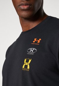 Crna pamučna majica s višestrukim logotipima: narančasti logotip Under Armour, okrugli logotip i žuti detalj brendiranja, okrugli izrez.