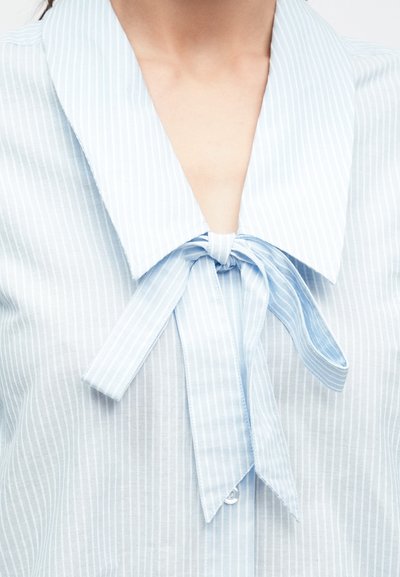 Svetlomodrá blúzka s prúžkami, ktorá má veľký golier s mašľou na krku, vyrobená z hladkej, ľahkej látky.