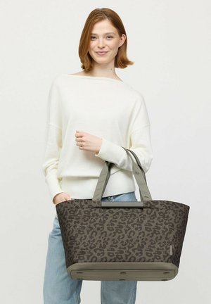 Junge Frau mit schulterlangem braunem Haar, die einen weißen Pullover und Jeans trägt und eine große graue Tasche mit Leopardenmuster hält.