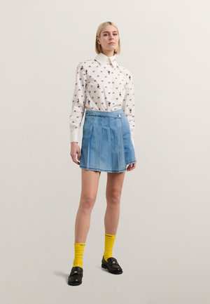 Hvit langermet skjorte med et lite blomstermønster, kombinert med en lys blå denimskjørt med folder. Svart loafers og gule knelengde sokker.
