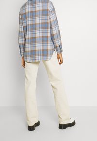 Persona che indossa una camicia a quadri blu e marrone, pantaloni crema e stivali bianchi con suole nere, in piedi contro uno sfondo chiaro uniforme.