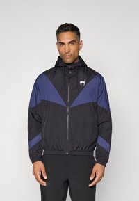 Veste coupe-vent noire et marine avec capuche. Présente une fermeture éclair à l'avant et des poignets élastiques, avec un logo sur la poitrine et un motif en chevrons.