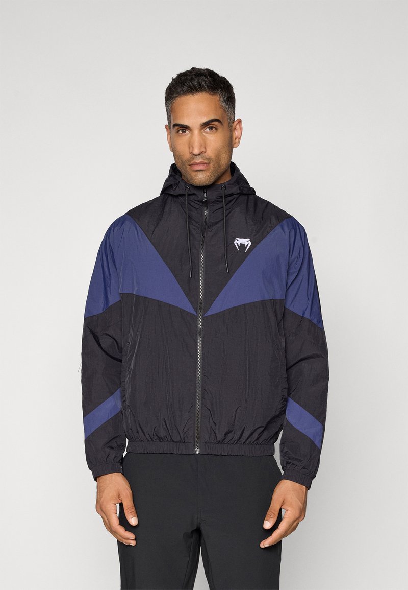 Veste coupe-vent noire et marine avec capuche. Présente une fermeture éclair à l'avant et des poignets élastiques, avec un logo sur la poitrine et un motif en chevrons.
