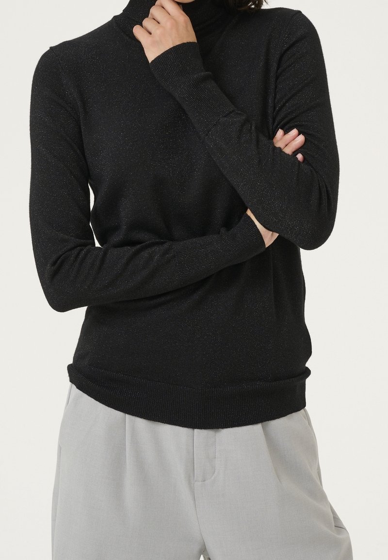 Personne portant un pull à col roulé noir à manches longues et un pantalon gris clair, les bras croisés et une main près du menton.