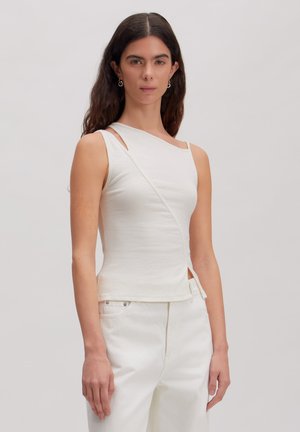 Femme aux longs cheveux foncés portant un haut asymétrique blanc sans manches et un pantalon blanc, se tenant devant un fond gris clair.