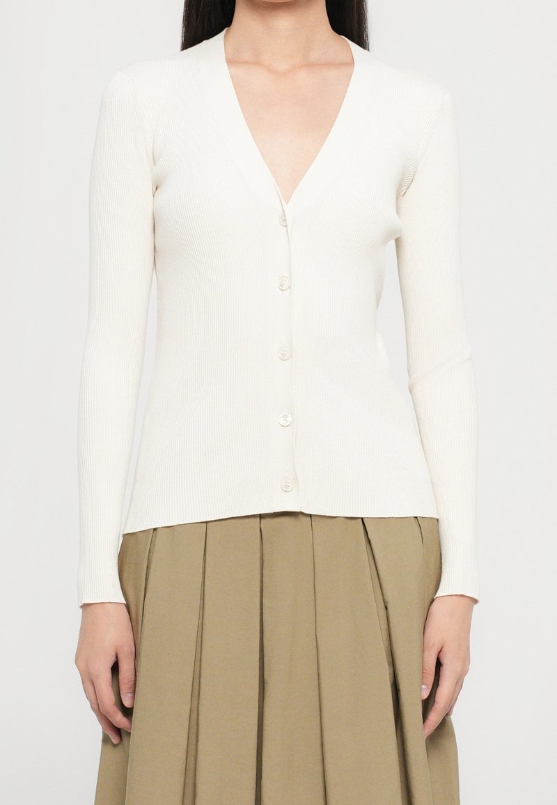 Femme portant un cardigan blanc côtelé à manches longues avec col en V et boutons, et une jupe plissée beige sur un fond uni.