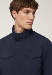 Marineblauwe waterdichte jas met een hoge kraag, twee borstzakken en een gestructureerde stof. Heeft subtiele branding op de linkerzak.