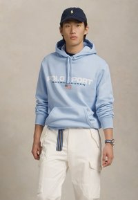 Γαλάζια hoodies με το κείμενο "POLO SPORT RALPH LAUREN" και αμερικανική σημαία, συνδυασμένο με λευκά cargo παντελόνια που διαθέτουν ριγέ ζώνη.
