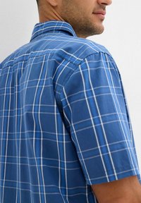 Camicia a maniche corte in cotone blu con un motivo a quadri blu e bianchi. Presenta un colletto classico e dettagli di cucitura visibili.