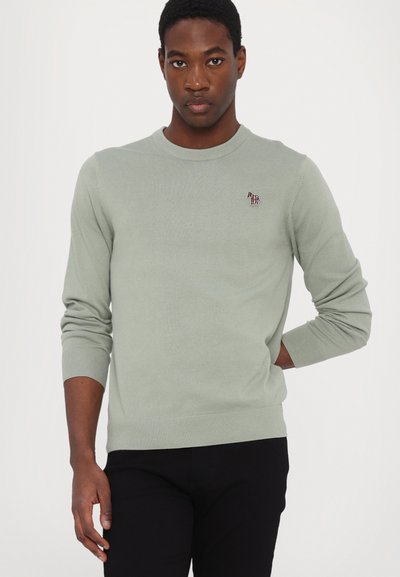 Suéter verde claro com decote redondo, punhos e bainha canelados, apresentando um pequeno logótipo de cavalo multicolorido no peito. Usado com calças pretas.