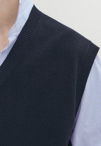 Mörk stickad väst med ribbat mönster, v-ringning, bärs över en ljusblå randig skjorta, med en ren, strukturerad design.