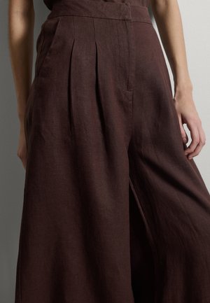 Pantalones de lino marrón oscuro de tiro alto y pierna ancha, con pliegues delanteros y bolsillos laterales, usados por una persona con las manos parcialmente visibles.