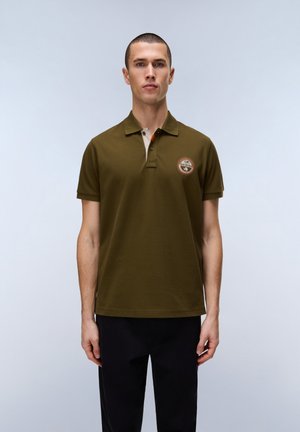 Młody mężczyzna w oliwkowej zielonej koszulce polo z okrągłą naszywką na lewej piersi, stojący na jednolitym jasnoszarym tle.