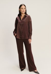 Camisa de satén marrón con botones y mangas acampanadas, combinada con pantalones anchos marrones a juego. La modelo lleva unos tacones negros de punta.