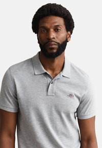 Homme barbu portant un polo gris clair à manches courtes avec un petit logo brodé sur la poitrine, faisant face vers l'avant.