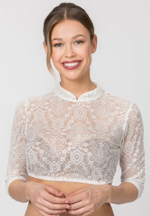 Lächelnde Frau mit hellbraunen Haaren trägt ein weißes, florales Spitzen-Cropped-Top mit dreiviertel Ärmeln und kleinem Stehkragen.