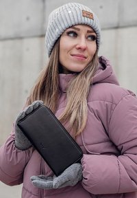 Black leather wallet held in grey gloves. The person wears a light purple puffer jacket and a grey and white striped beanie. Background is concrete.