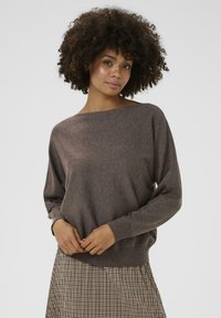 Brūns, mīkstajā adījumā izstrādāts džemperis ar plašu kuģa kakla griezumu un garām piedurknēm. Riboti aproces un apakša. Nēsāts virs raibā svārku raksta.