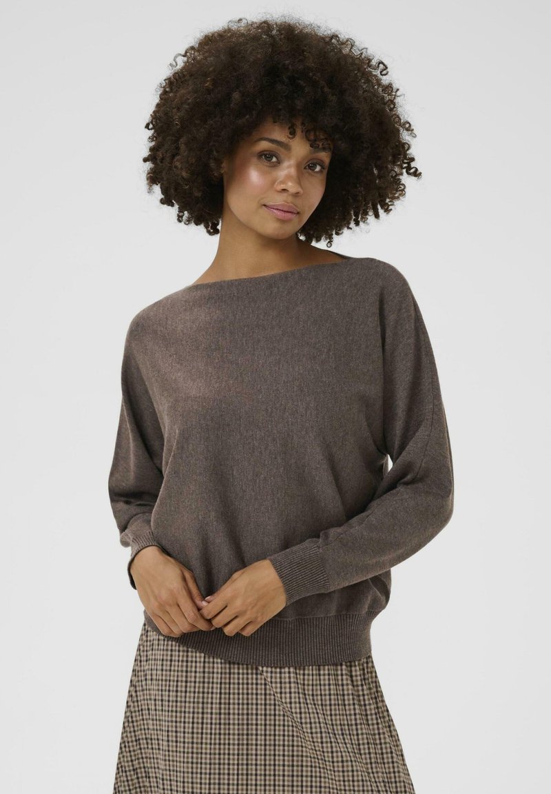 Brūns, mīkstajā adījumā izstrādāts džemperis ar plašu kuģa kakla griezumu un garām piedurknēm. Riboti aproces un apakša. Nēsāts virs raibā svārku raksta.