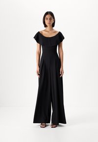 Melna ķermenis bez plecēm ar plisētām platām kājām. Gluda auduma struktūra rada plūstošu siluetu. Valkāts ar melniem sandaļiem.