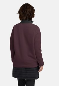 Bordeaux fleece jack met een hoge kraag, gecombineerd met een zwarte puffer onderlaag. Heeft lange mouwen, een ontspannen pasvorm en een gladde textuur.