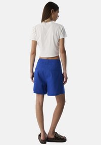 T-shirt blanc cropped associé à un short bleu, présentant une texture lisse et deux poches arrière. Porté avec des sandales sombres.