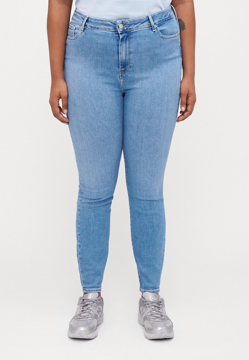 Jeans de mezclilla azul claro de cintura alta, corte ajustado, diseño de cinco bolsillos y sutiles desgastes. Combinados con zapatillas gris.