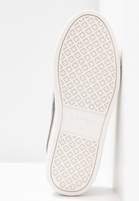 Semelle de chaussure blanche avec motif de rainure en losange et "Esprit 37" embossé au centre, positionnée contre un mur d’angle blanc.