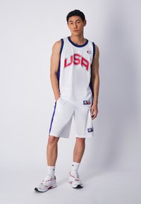 Camisa de baloncesto blanca con "USA" en texto rojo, acompañada de pantalones cortos blancos a juego con acentos laterales azules, usados con zapatillas deportivas blancas.