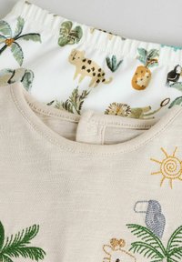 Beige shirt met geborduurde dieren en zon, gecombineerd met witte shorts met kleurrijke jungle-dierafdrukken en palmbladeren.