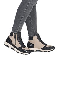 Baskets montantes noires et brunes avec un dessus en daim et cuir verni, dotées d'une fermeture éclair sur le côté et d'une semelle en caoutchouc contrastante.