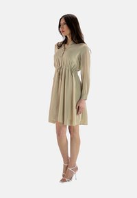 Beige, langärmliges Kleid mit einer Kordelzug-Taille und knöpfbarem Ausschnitt. Fließender Stoff mit einem gerafften Rock; kombiniert mit weißen Riemchen-Heels.