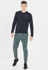 Schwarz langärmliges Sportshirt mit einem dezenten Logo, kombiniert mit hellgrünen Hosen, die schwarze seitliche Akzente haben. Schwarze Sportschuhe.