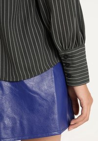 Schwarz gestreiftes Langarmshirt mit Knopfmanschetten, kombiniert mit einem glänzenden blauen Lederrock, der kontrastreiche Texturen und Farben betont.